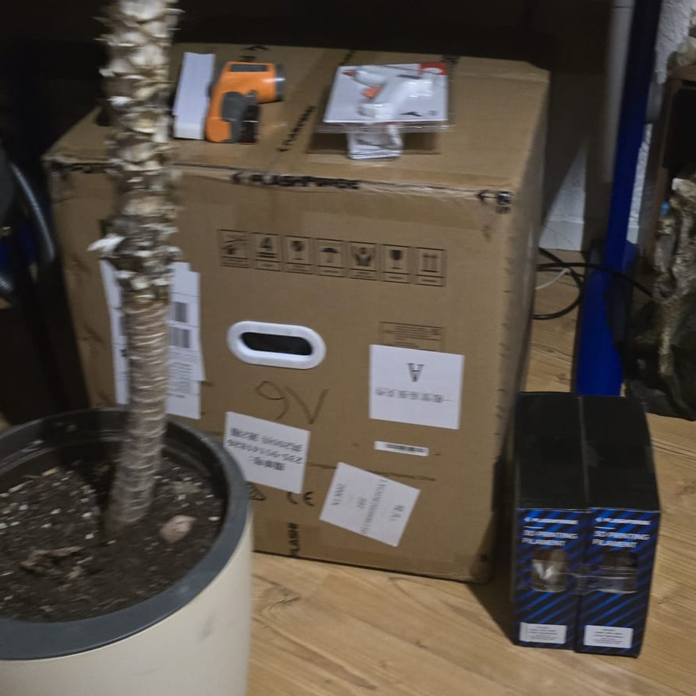 Der noch verschlossene FlashForge-Karton auf dem Boden, daneben zwei Filamentboxen, obendrauf ein Infrarot-Thermometer und eine Klebepistole