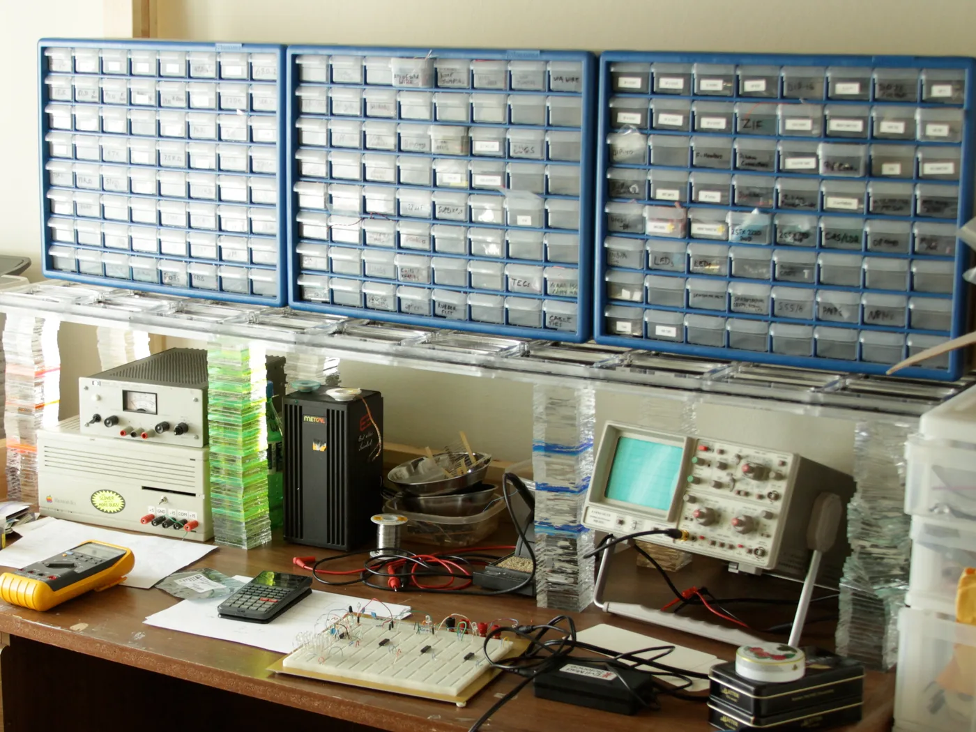 Elektronik-Arbeitsplatz mit drei Wänden aus beschrifteten Sortierschubladen, Oszilloskop, Labornetzteil, Multimeter und Steckbrett