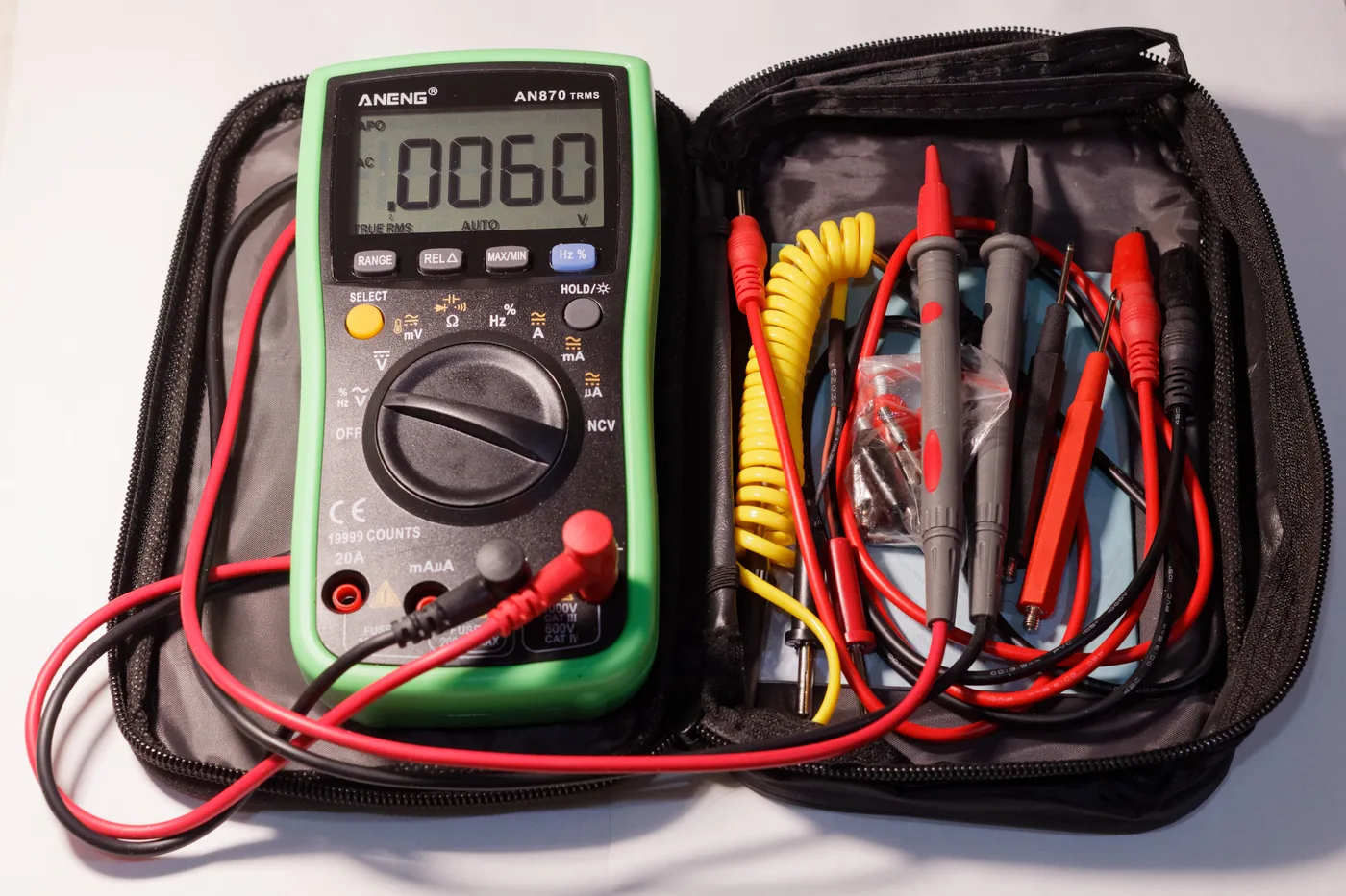 Digitales Multimeter mit grüner Schutzhülle in einer geöffneten Tasche, daneben Messleitungen, Prüfspitzen und Krokodilklemmen