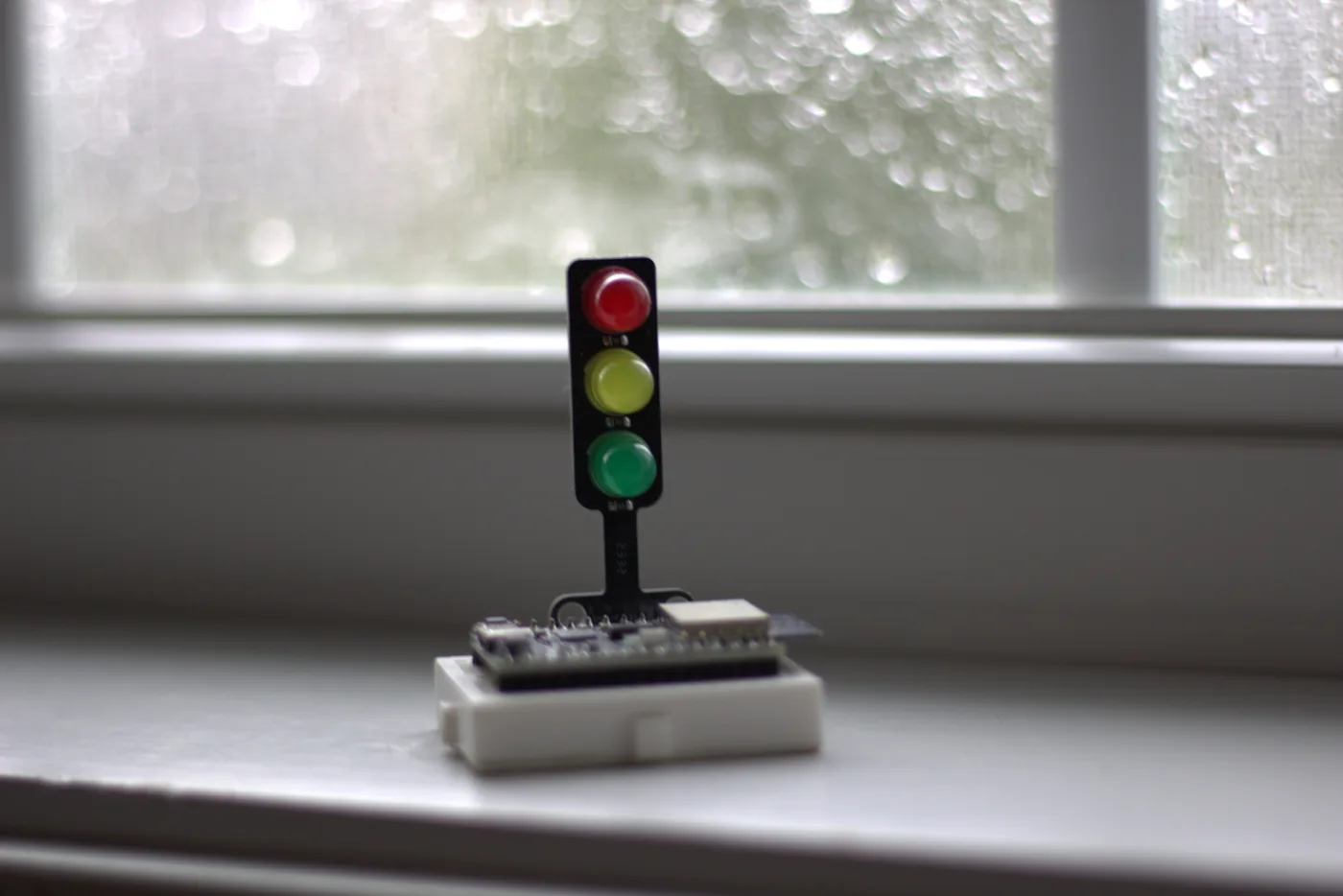 Kleines Mikrocontroller-Board auf einem weißen Steckbrett, darauf ein Ampel-Modul mit roter, gelber und grüner LED, vor einem regennassen Fenster