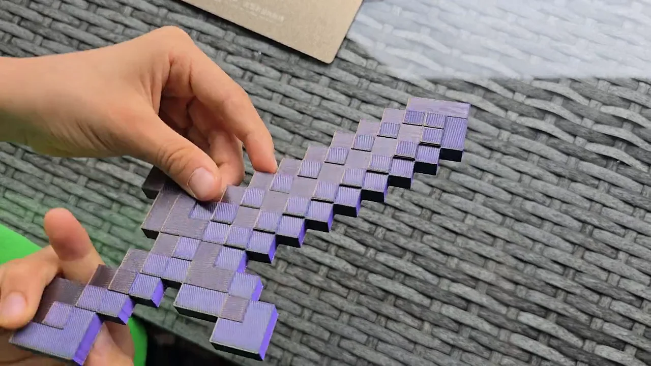 Zwei Hände halten ein gedrucktes Pixel-Schwert aus lila schimmerndem Filament über einem Rattantisch, an den Kanten sind die Druckschichten zu sehen